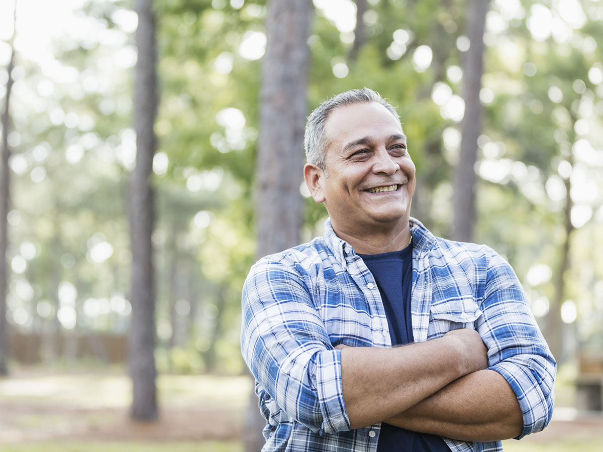 A mature man wearing a plaid shirt, standing in a park, smiling with his arms crossed.