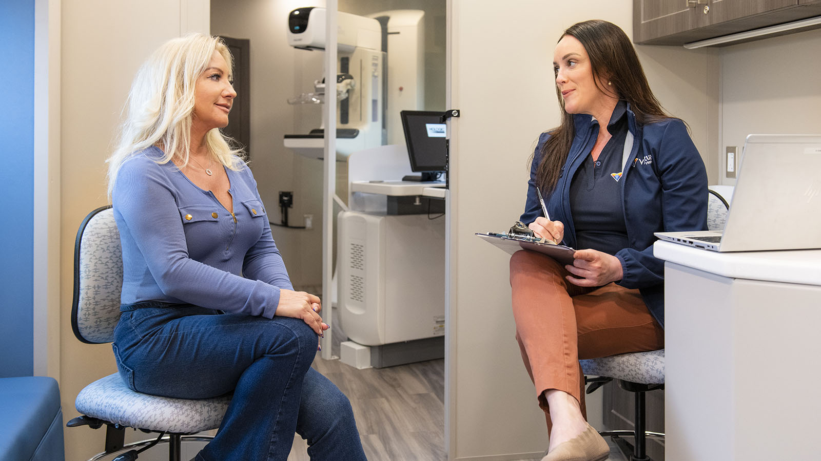 Woman at a computer with a clipboard talks to another woman seated in a chair in a mobile medical office