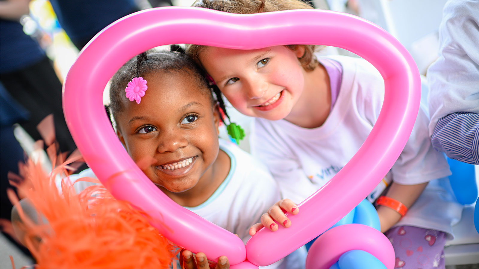 Two young female children pose framed by a heart-shaped pink balloon