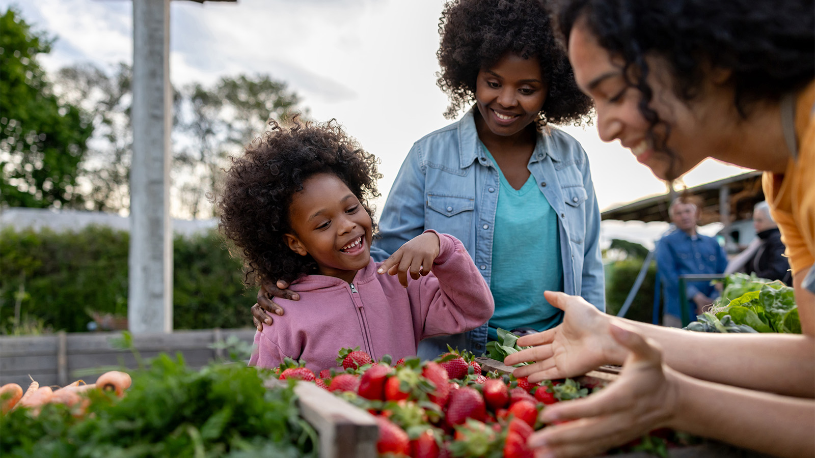 A mother and young daughter smile as another woman examines produce at a farmers market