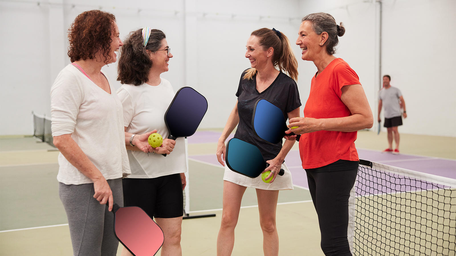 Group of 4 middle-aged women talking and laughing together on an indoor pickleball court