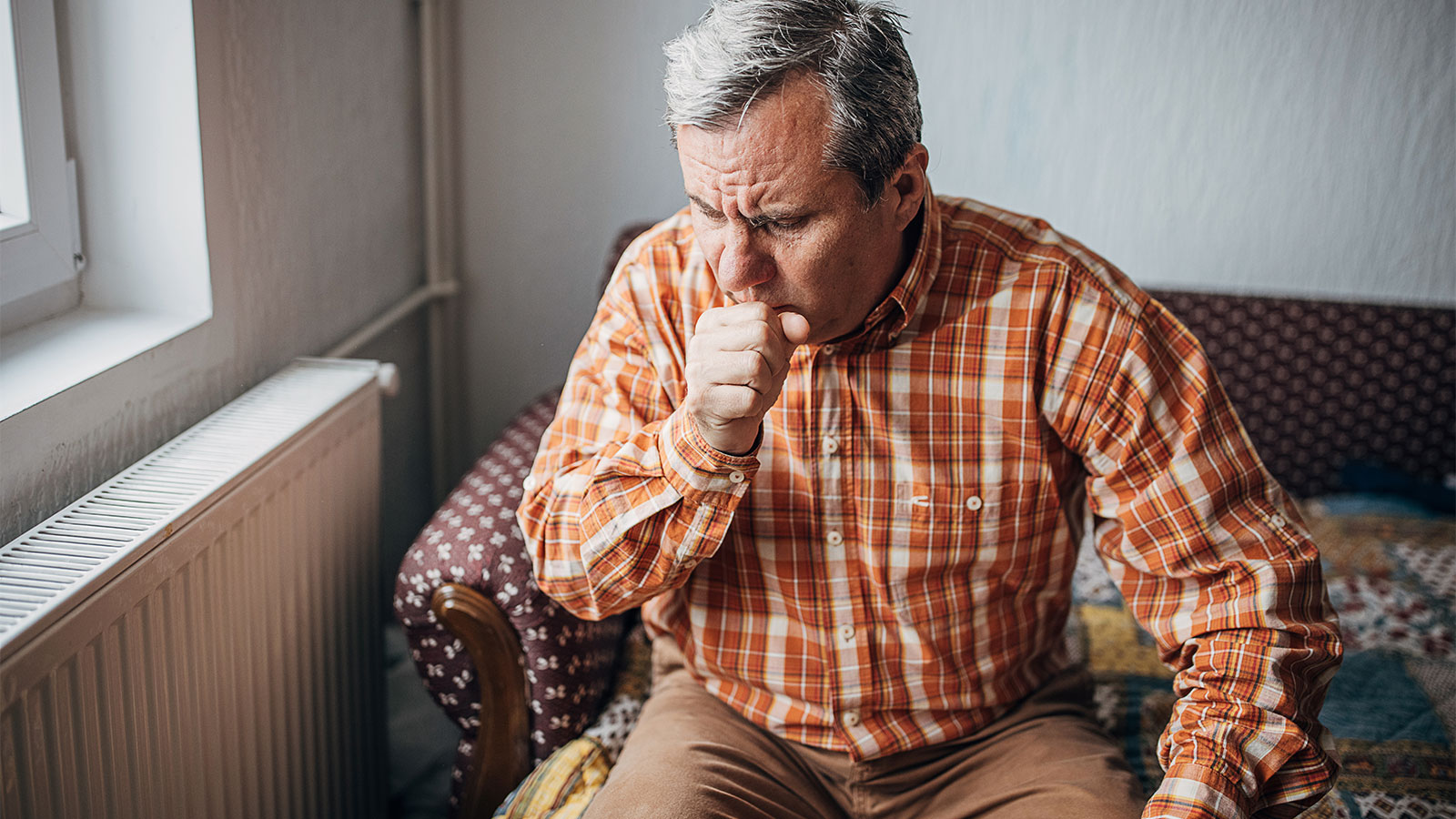 Male senior citizen sitting on couch, holding hand to mouth and coughing