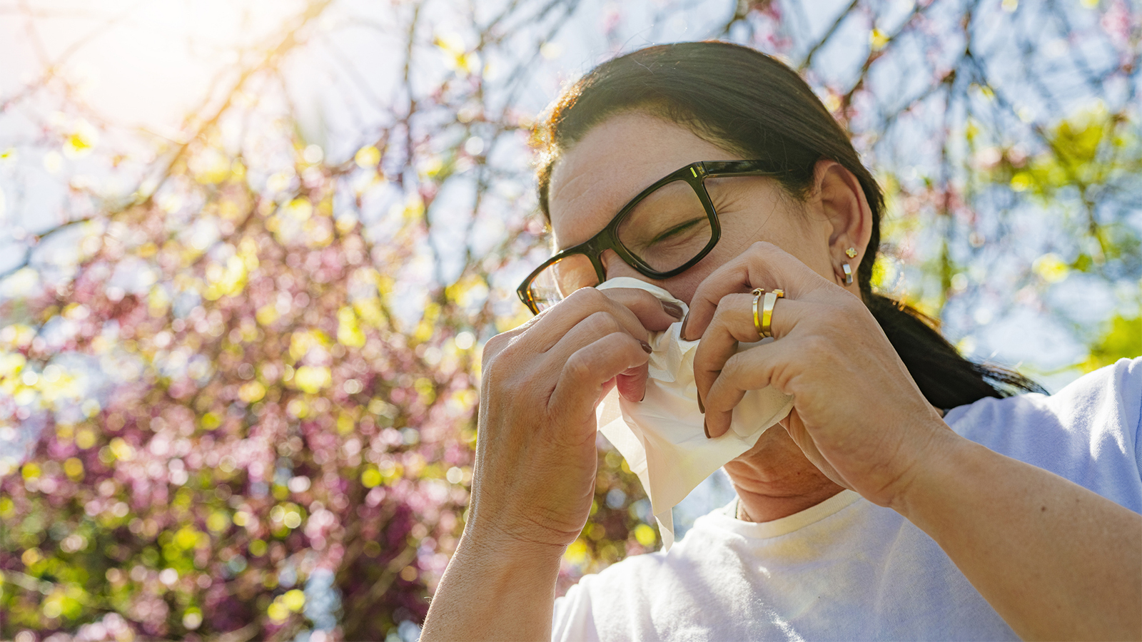 Woman wearing glasses, outside in nature, holding a tissue to her nose and sneezing