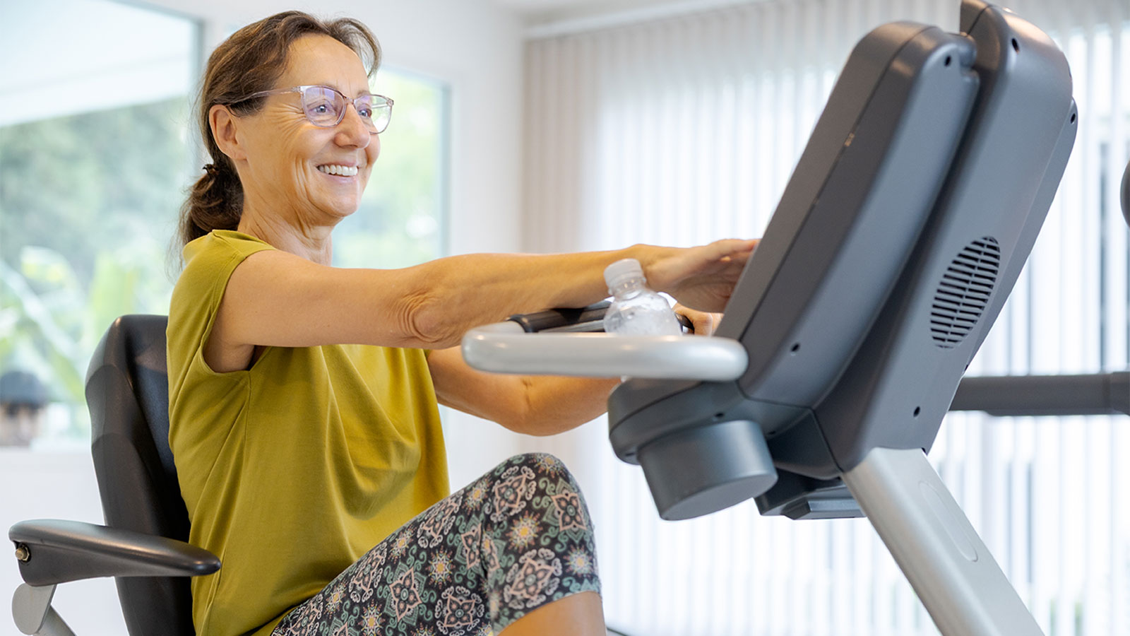 Woman with her hair in a ponytail and wearing glasses, smiles while touching the screen on a recumbent exercise machine.