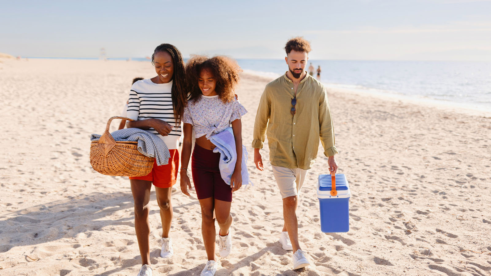 Two women and a man in their 20s, walking on the beach with a picnic basket, blankets and a cooler
