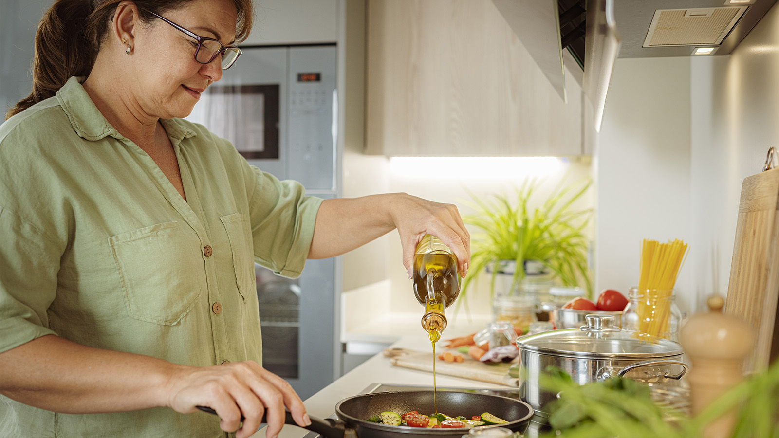 Woman pouring olive oil onto vegetables in a frying pan in her kitchen