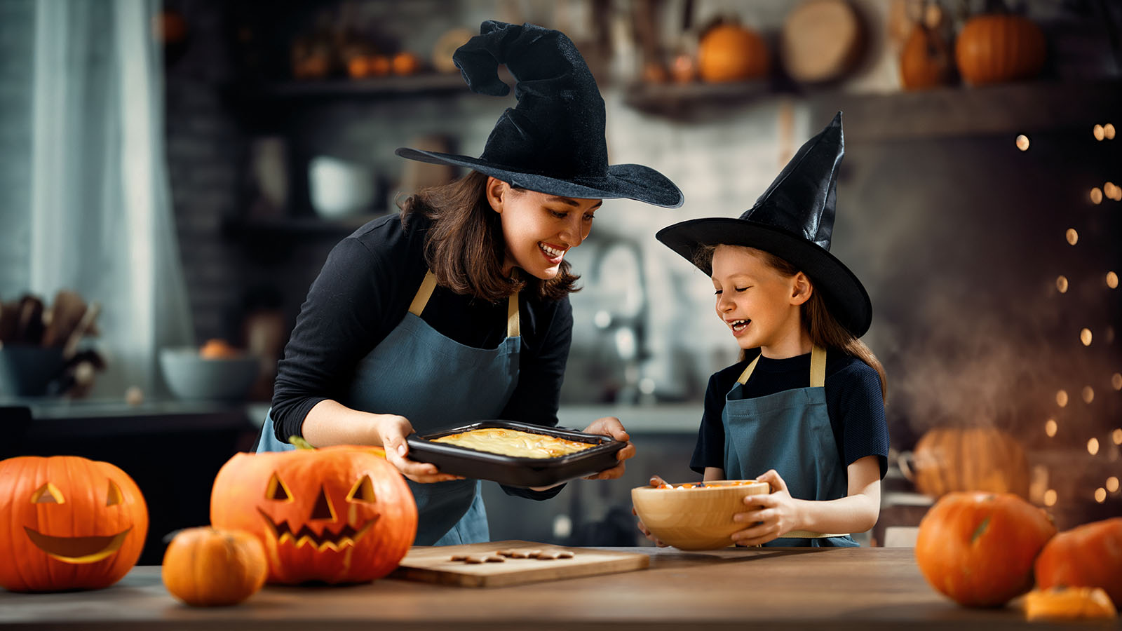 A woman and girl dressed in witch costumes stand in kitchen with Halloween decor