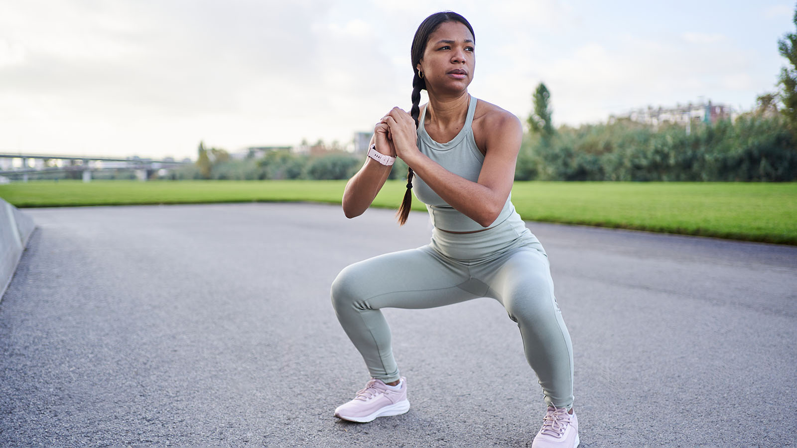 Woman in exercise outfit does squats on a paved track next to a grassy area