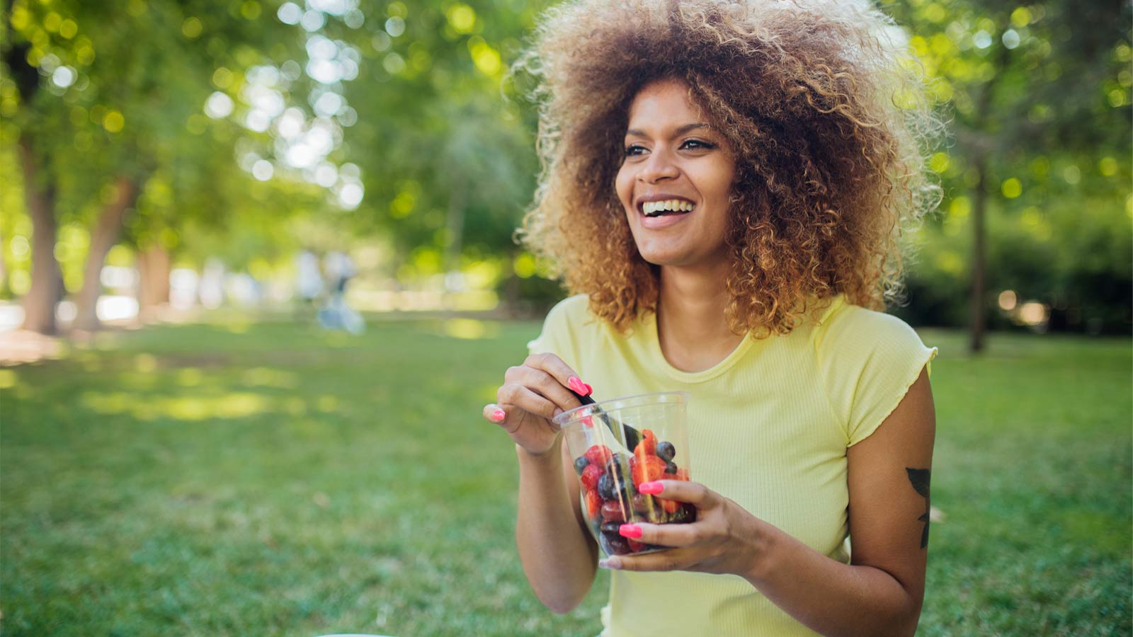 young woman sitting in a park eating a cup of fruit