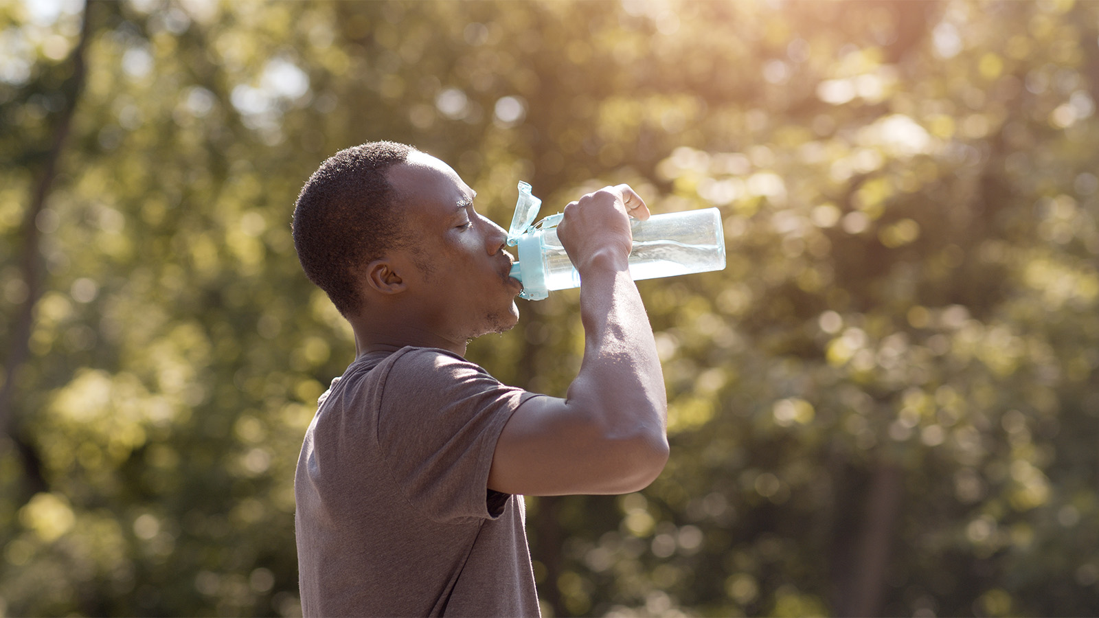 A male in his 30s drinks from a water bottle on a sunny day, with trees in the background
