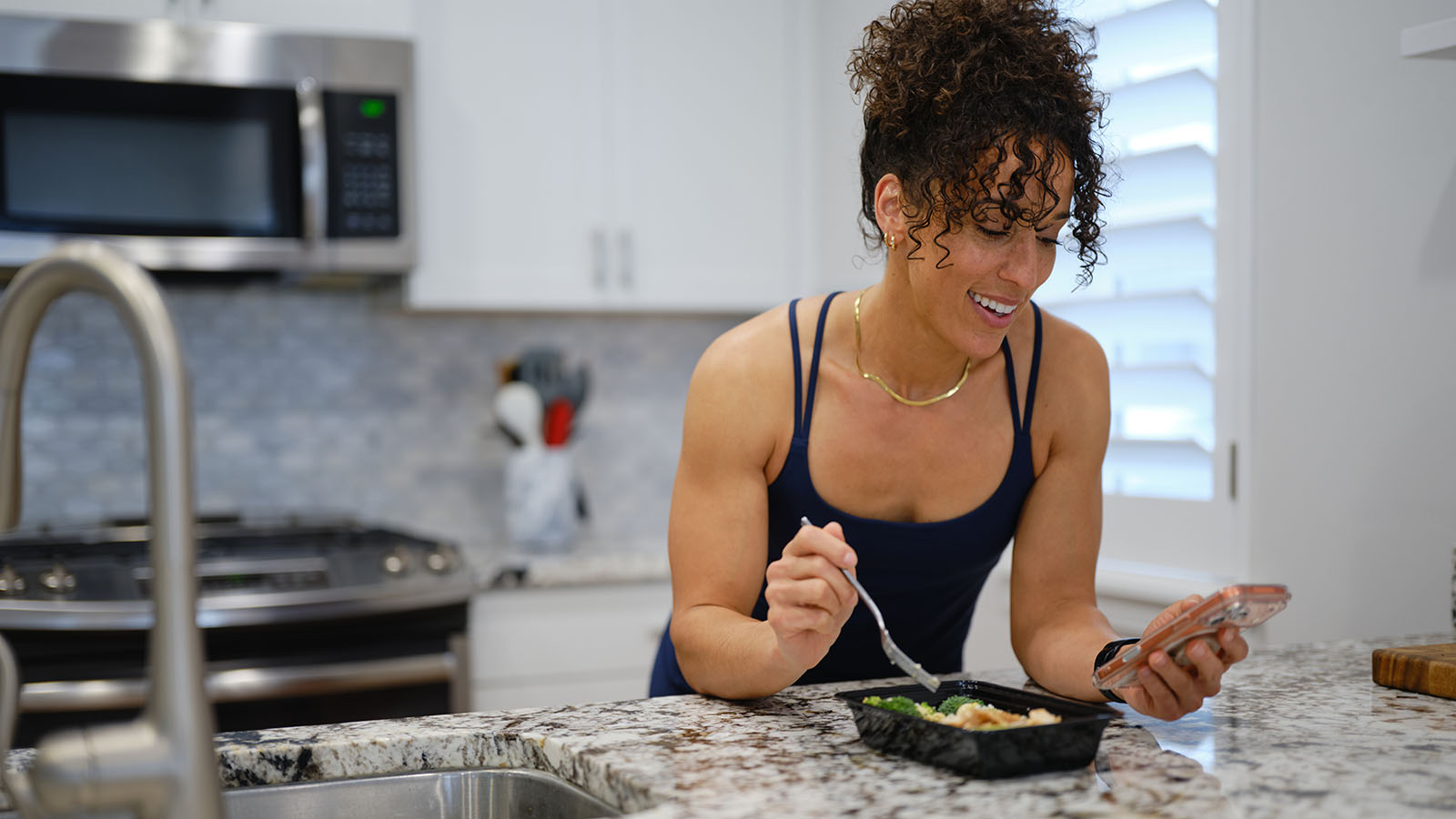 Woman eats a meal while smiling and holding cell phone in kitchen