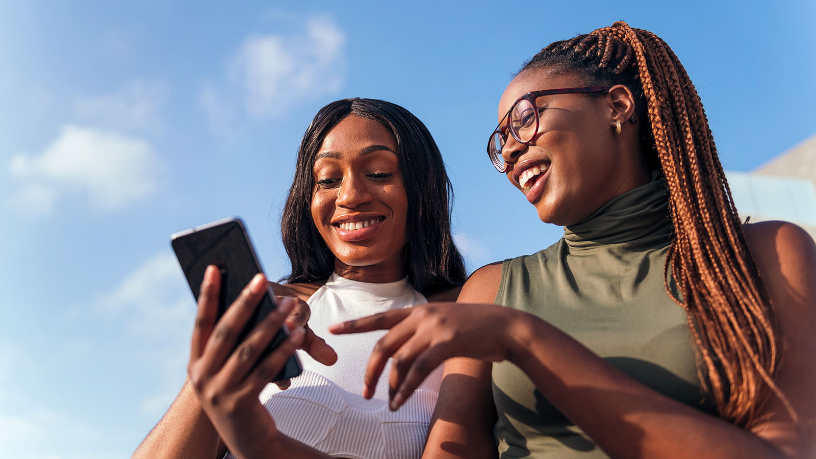 Two young women smiling and looking at a phone in an outdoor setting.