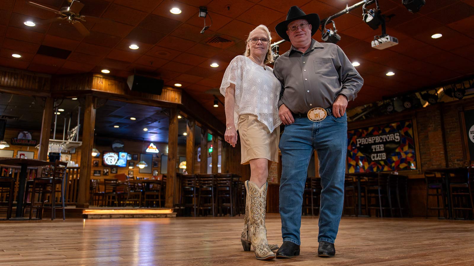 couple wearing cowboy boots and cowboy hats standing on a dancefloor in a bar
