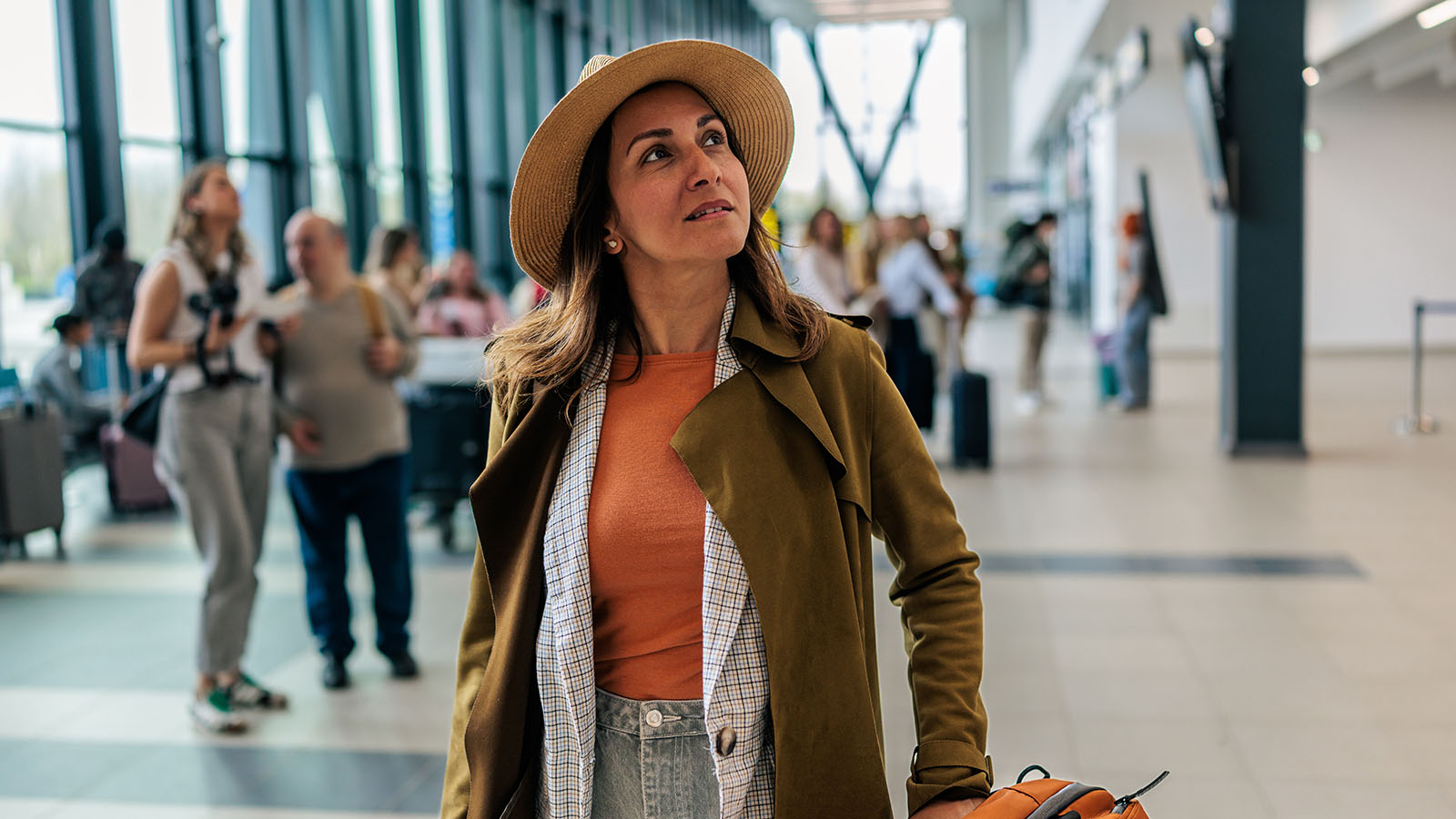 Woman in sunhat and brown coat pulling a suitcase in an airport with travelers in the background