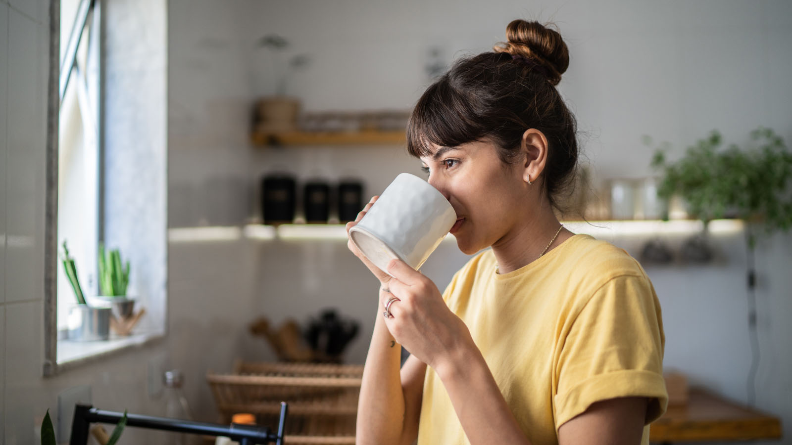 30-something woman in a yellow shirt, sipping from a coffee mug while standing in a kitchen