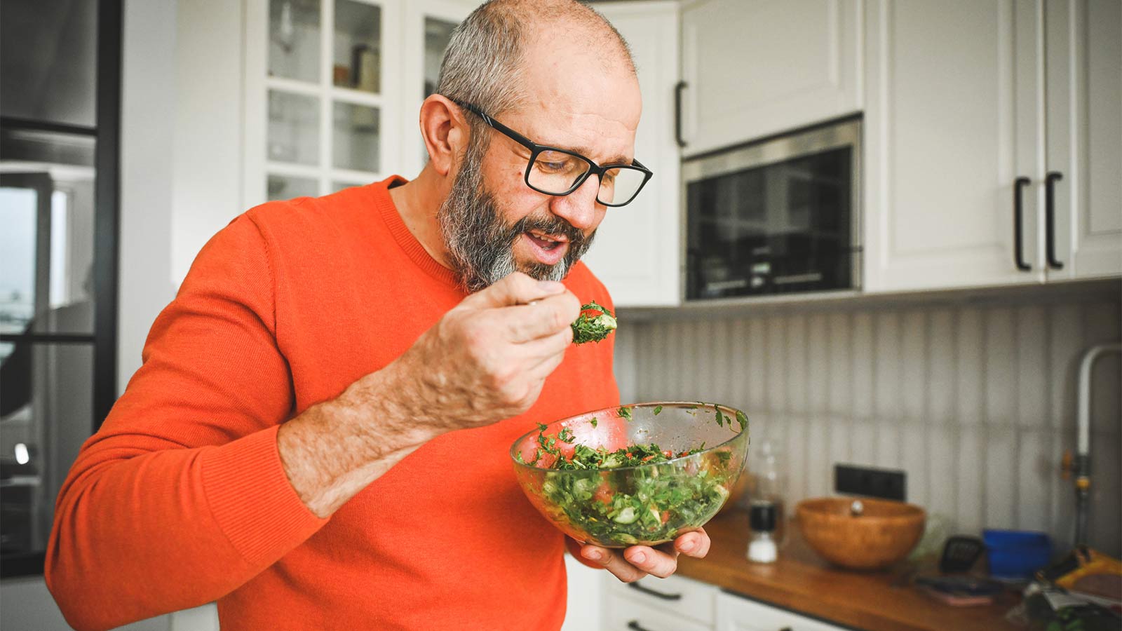 middle aged man standing in his kitchen eating salad