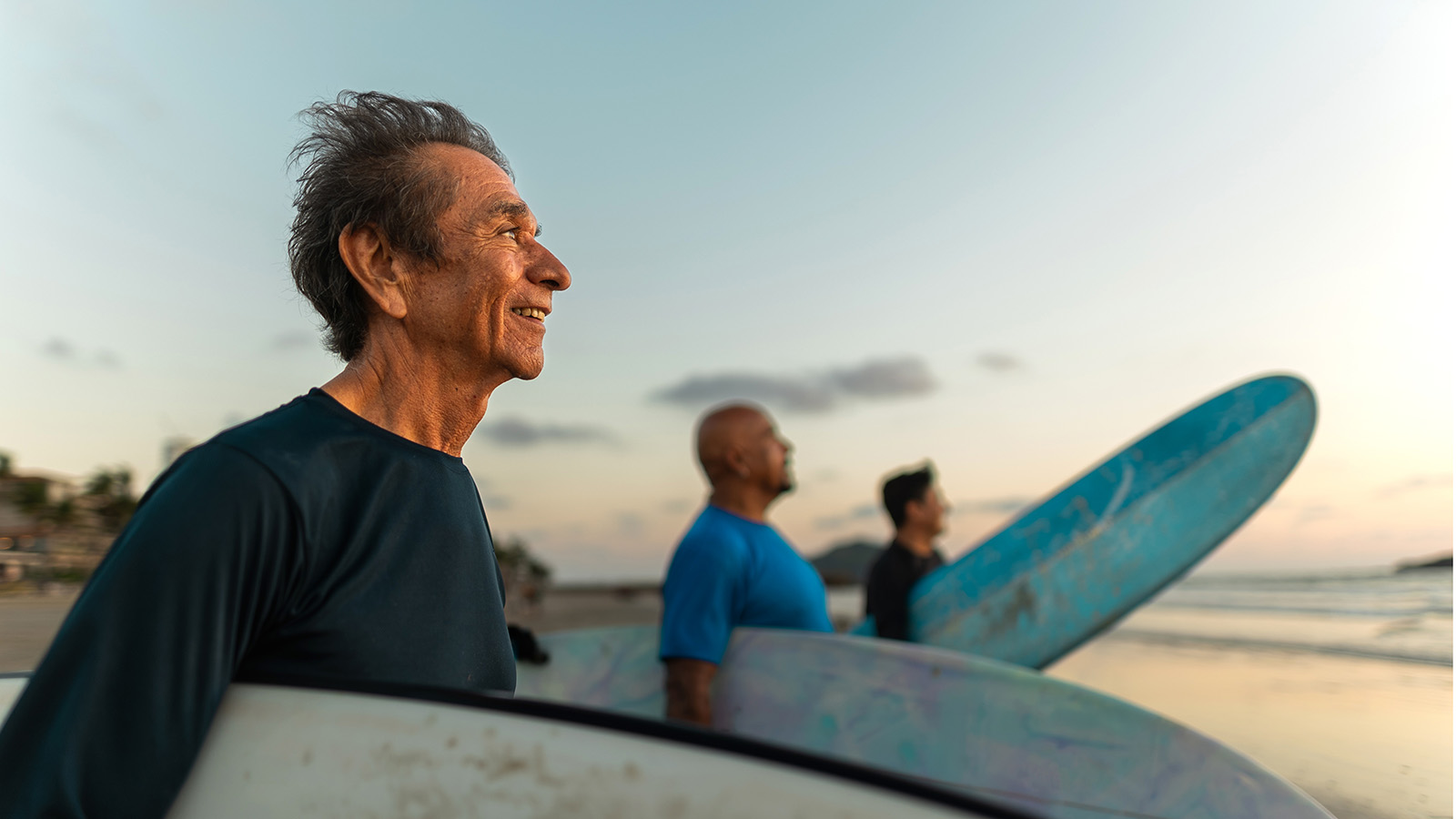 Older man and two other males all holding surg boards and looking at the ocean