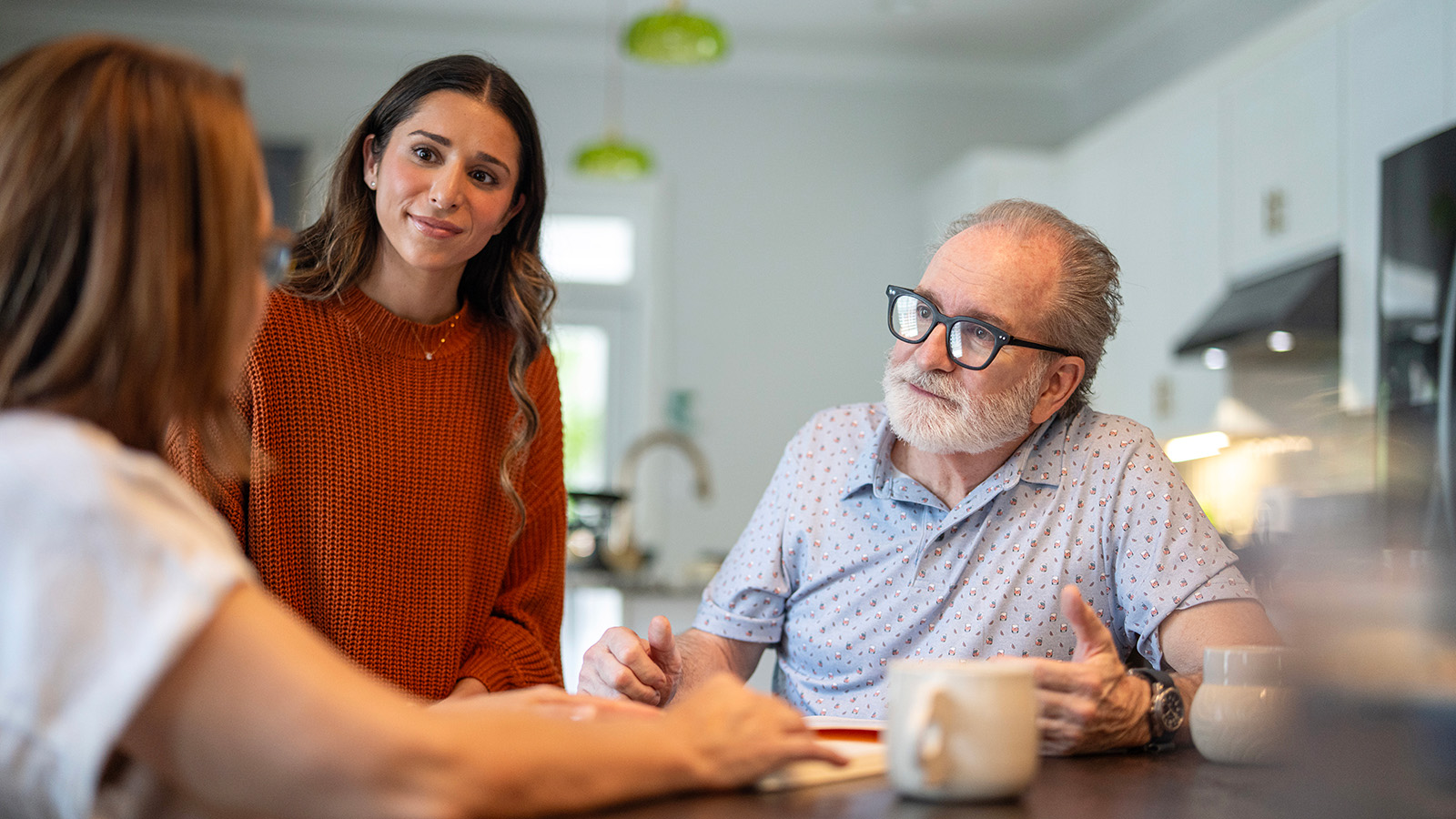 Young adult woman standing and talking with older adult couple seated at kitchen table