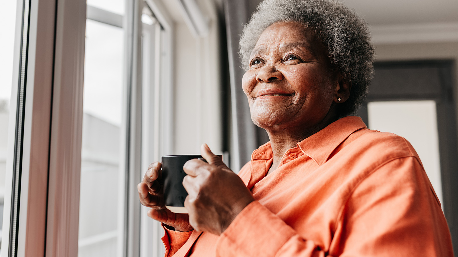 Older woman looking out a window and enjoying a cup of coffee