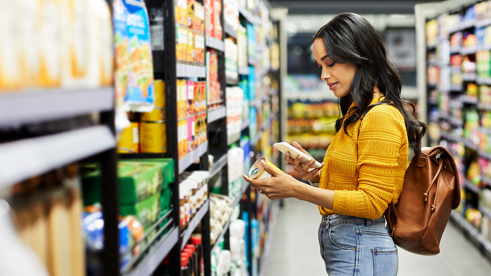 Young woman in grocery store aisle looks at phone while holding a canned good item