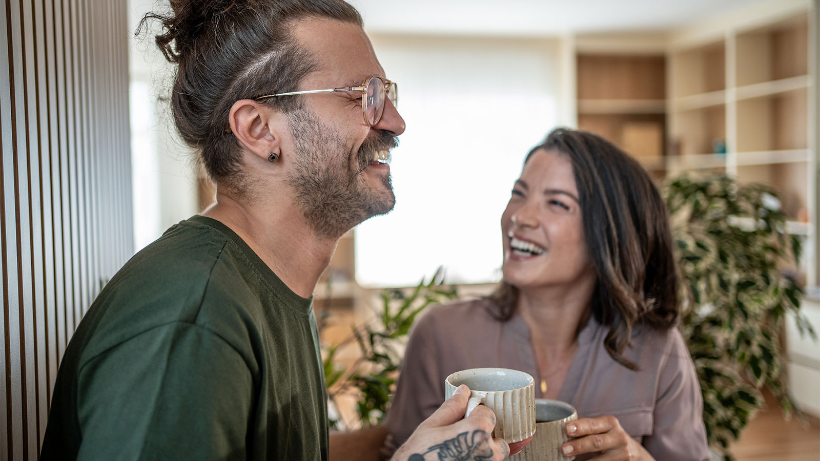 Man and woman holding coffee cups, smiling and laughing in their living room