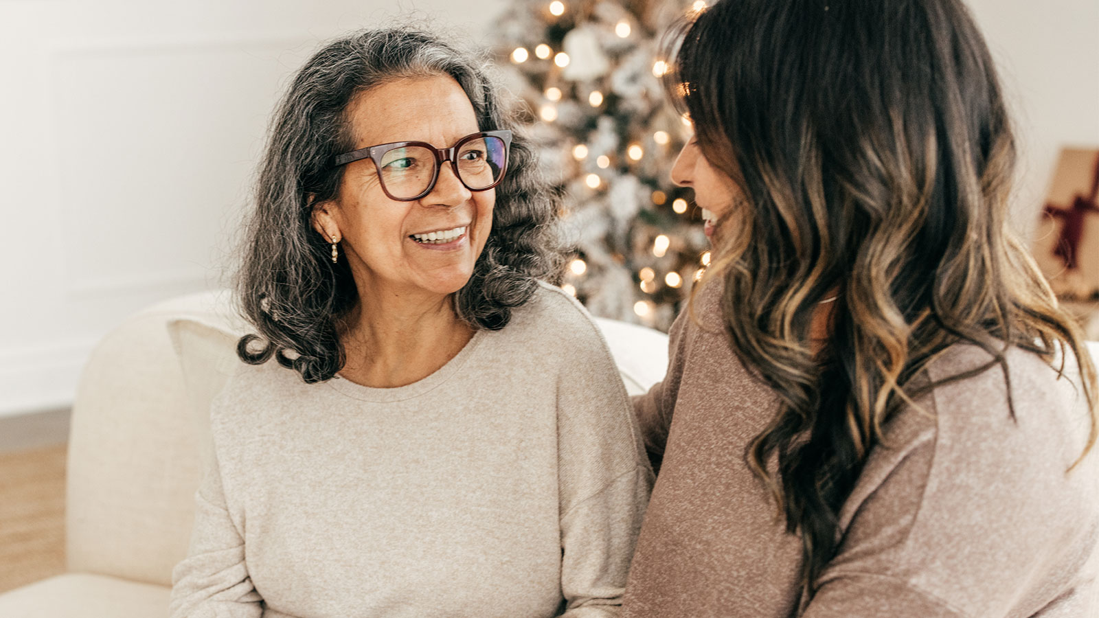 Older woman sitting on couch and smiling while talking to younger woman, with a Christmas tree in the background
