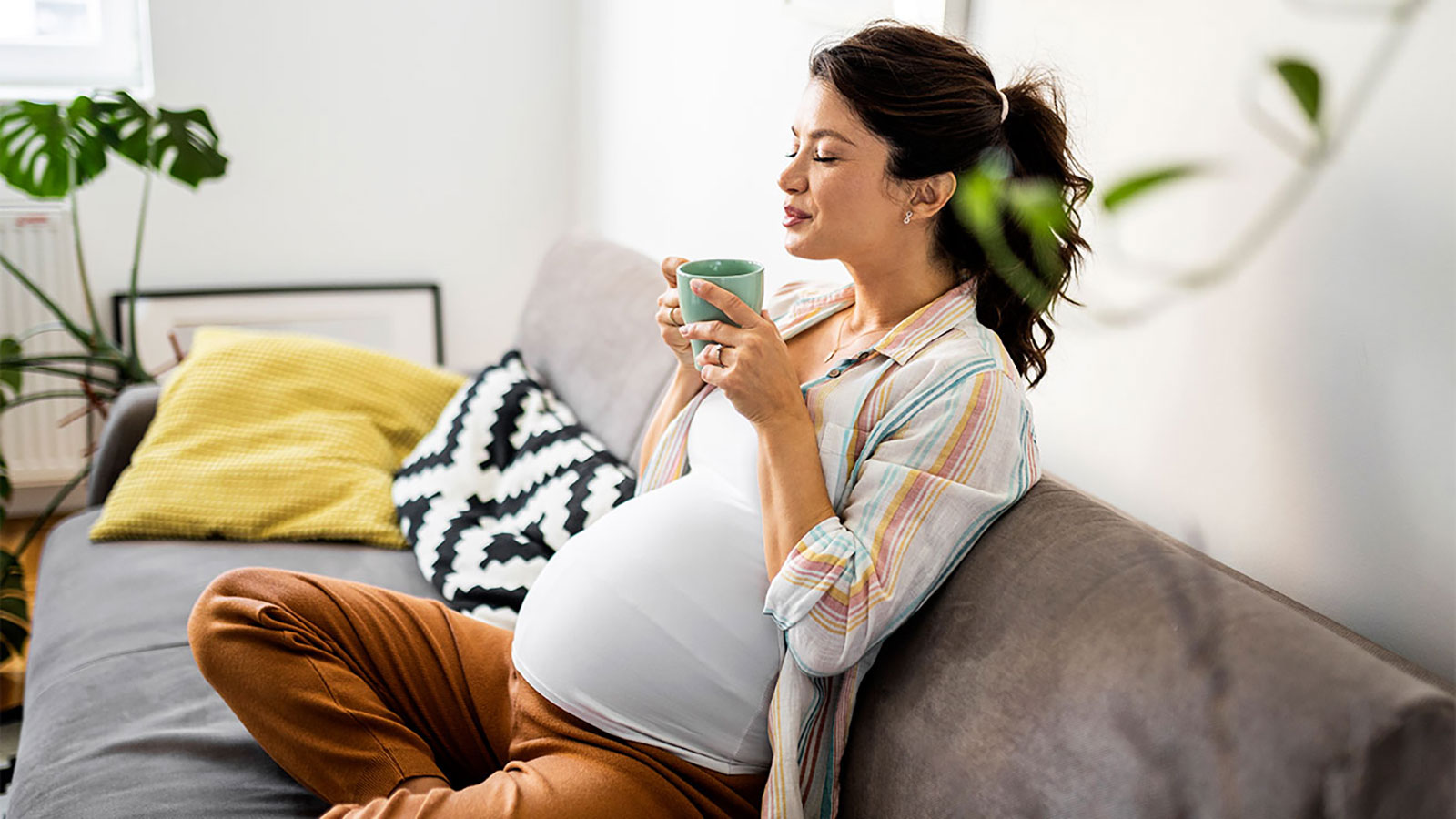 Pregnant woman with long hair in a ponytail relaxes on couch, holding a mug, smiling and with eyes closed