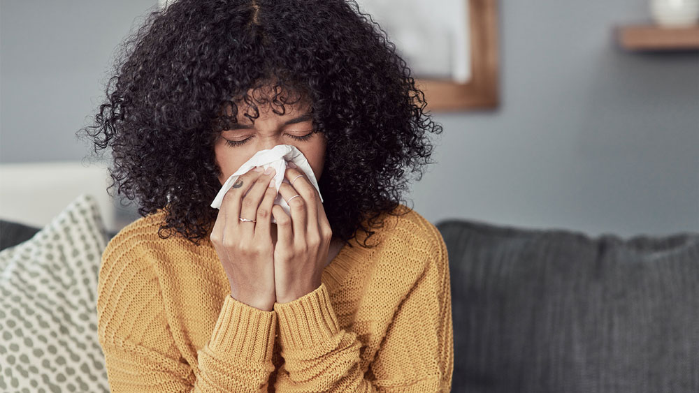 Woman wearing a yellow sweater, sitting on a couch and blowing her nose into a tissue