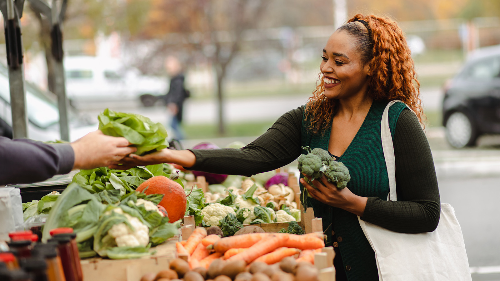 Woman at farm stand, smiling, holding broccoli and accepting lettuce from an unseen person at the stand
