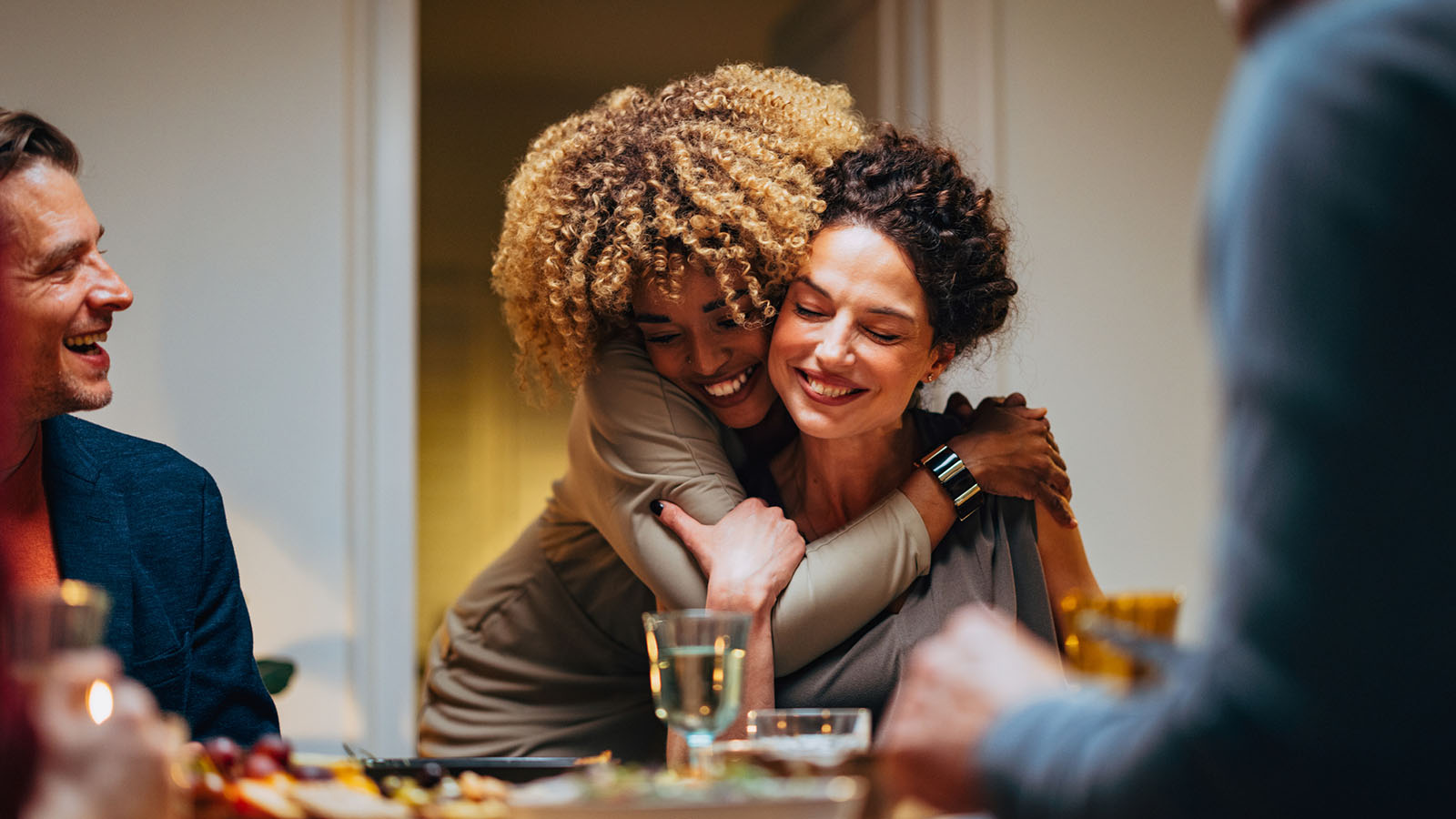 Woman is hugging her female friend from behind her chair; they are at a dinner table with friends and a glass of wine is on the table