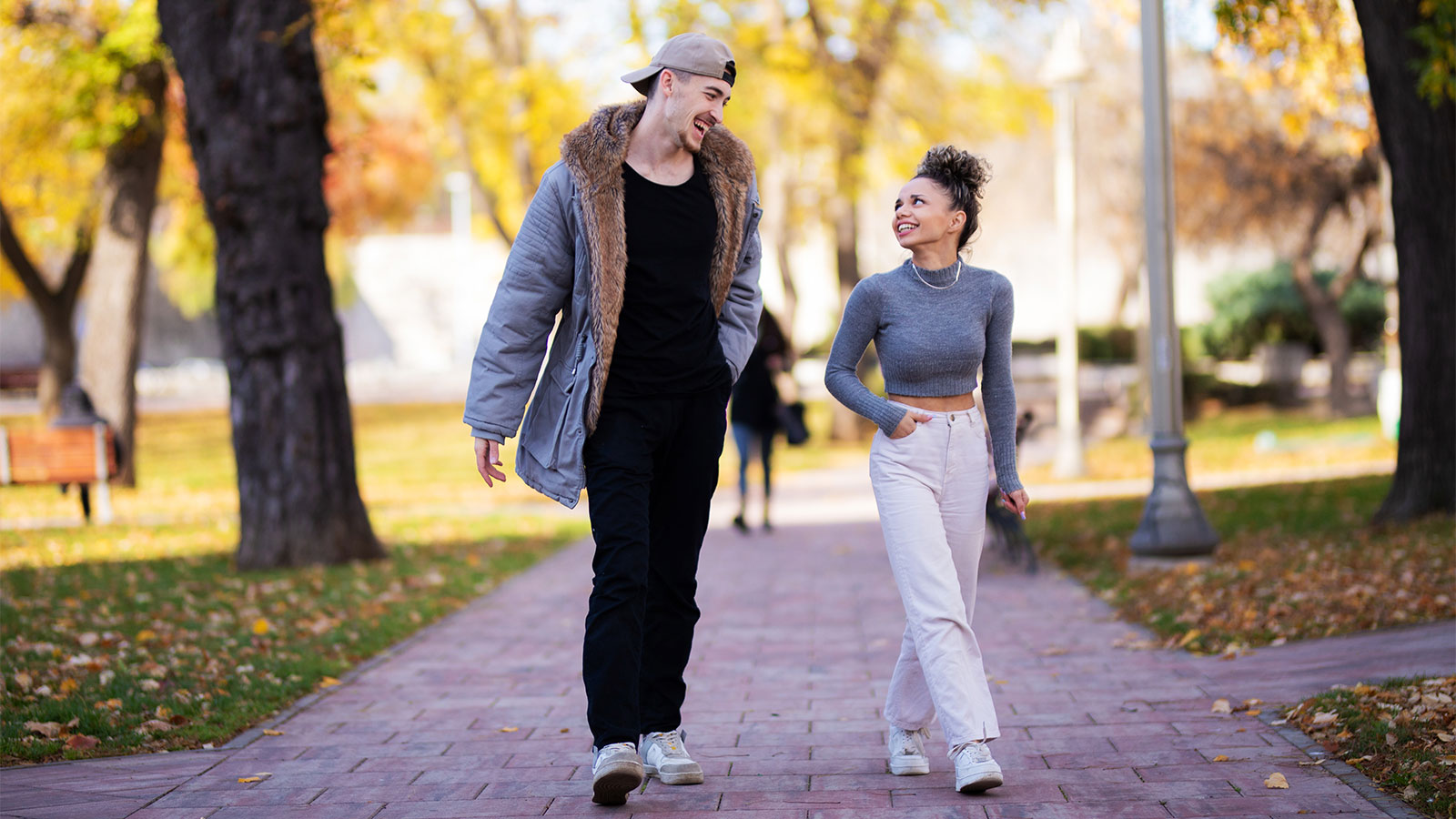 Young man and woman walking on a paved path. The path is lined with trees that have turned yellow in the fall.