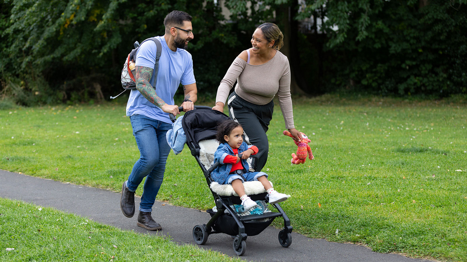 Mother and father talking and walking along a paved path in a park, with a toddler in a stroller
