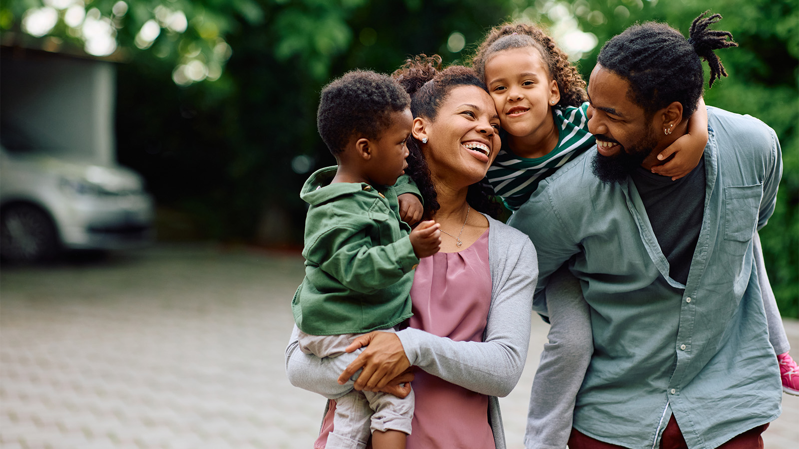 Mother holding male toddler, smiling at father who is giving female toddler a piggy-back ride, outdoors in their driveway