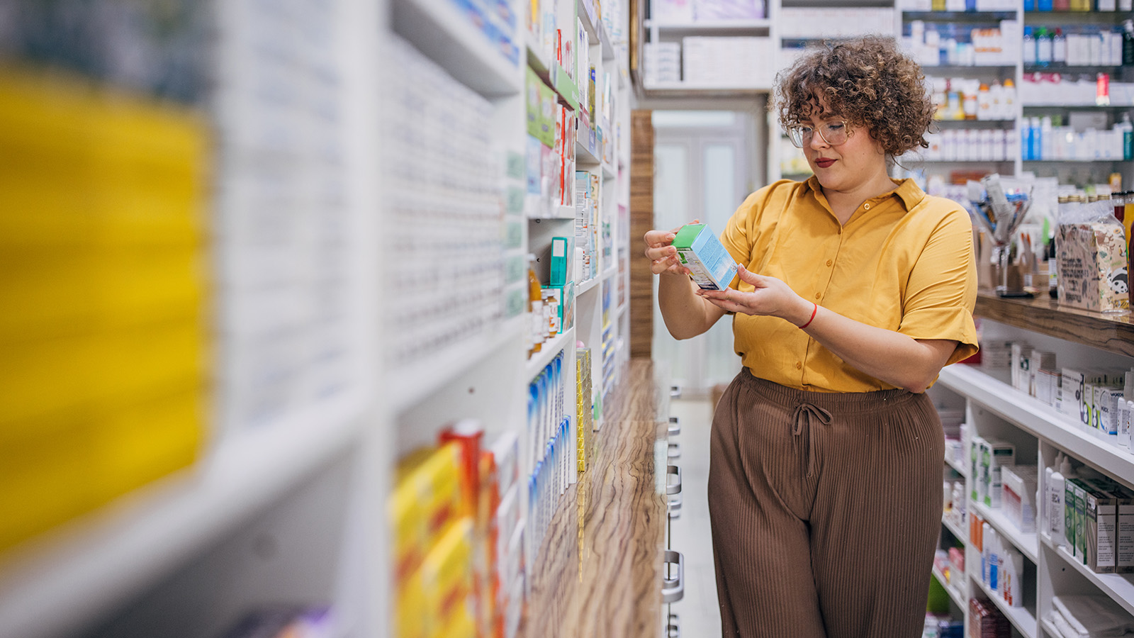 Woman in glasses holding a box of medication while standing among shelves at a drug store