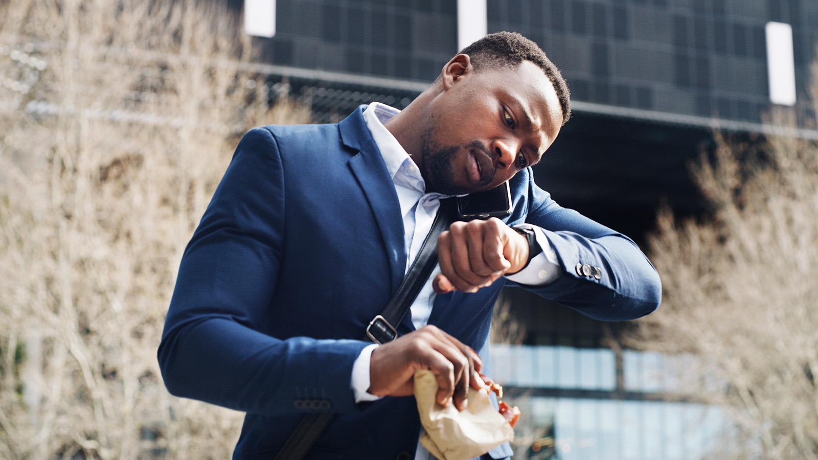 Young man in a suit, looking at his watch, walking and talking on the phone, while carrying a paper bag