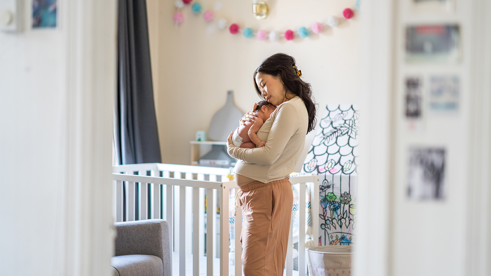 Woman with post-pregnancy belly holding newborn next to a crib in the baby's nursery