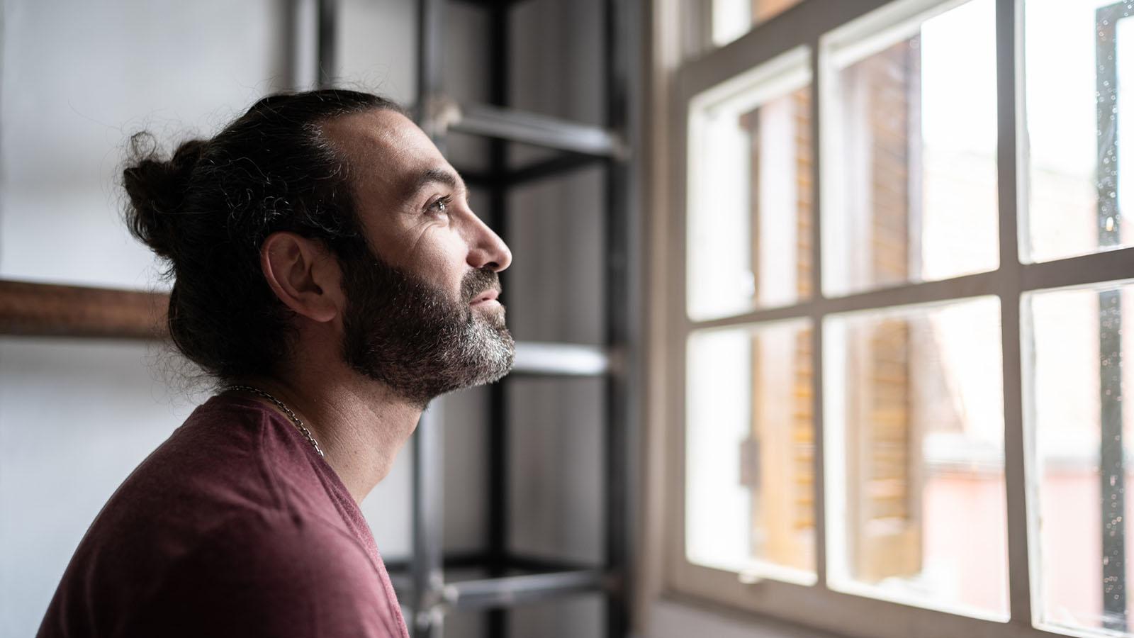 Man in his late 30s with hair in a bun, slightly smiling, looking out a window from inside an apartment