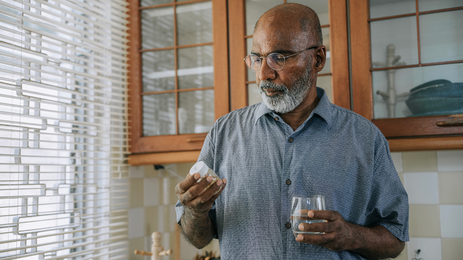 Older man examining pill bottle in one hand and holding a glass of water in his other hand