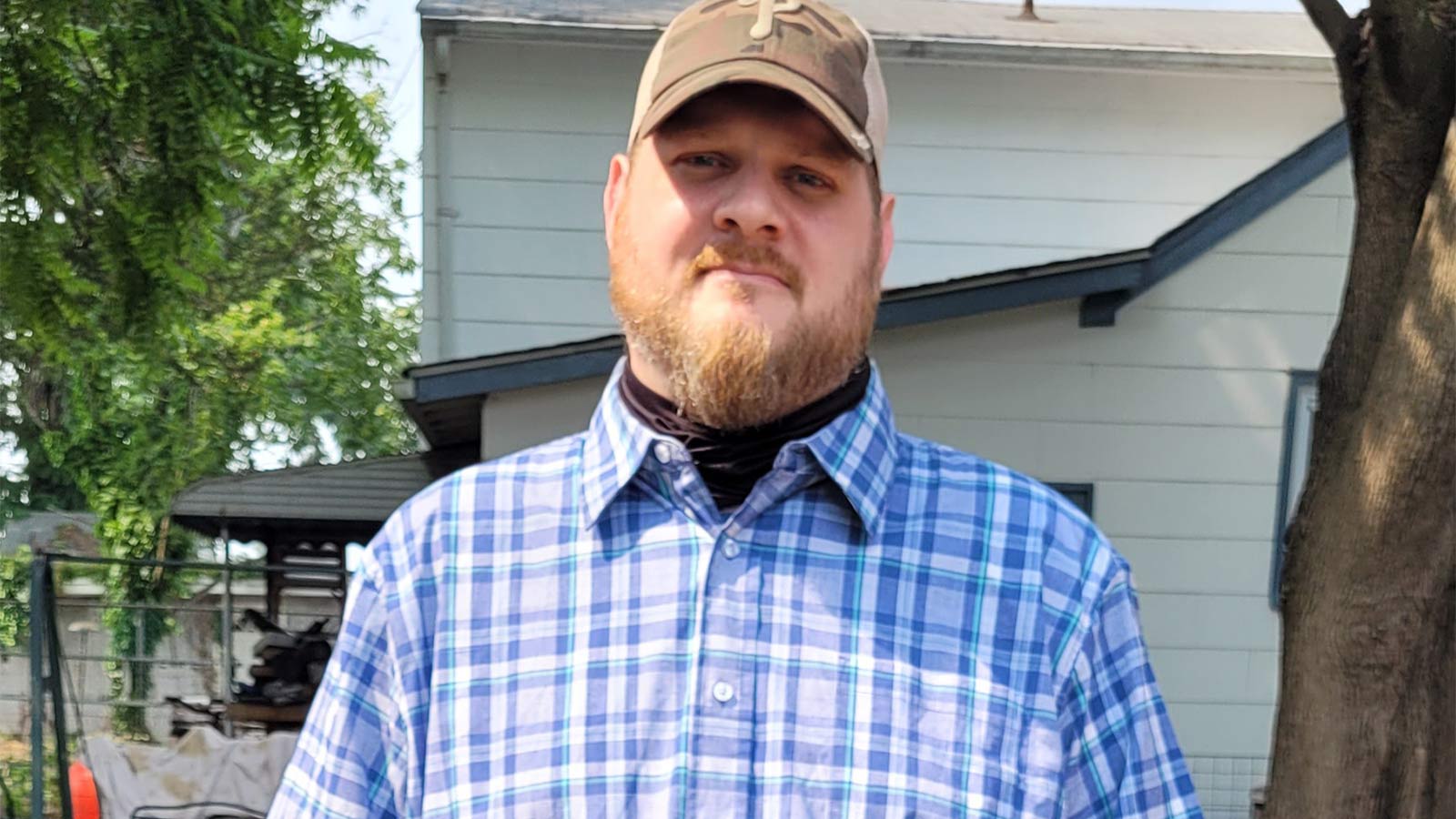 young man with beard in baseball cap and plaid shirt smiling outside of a house