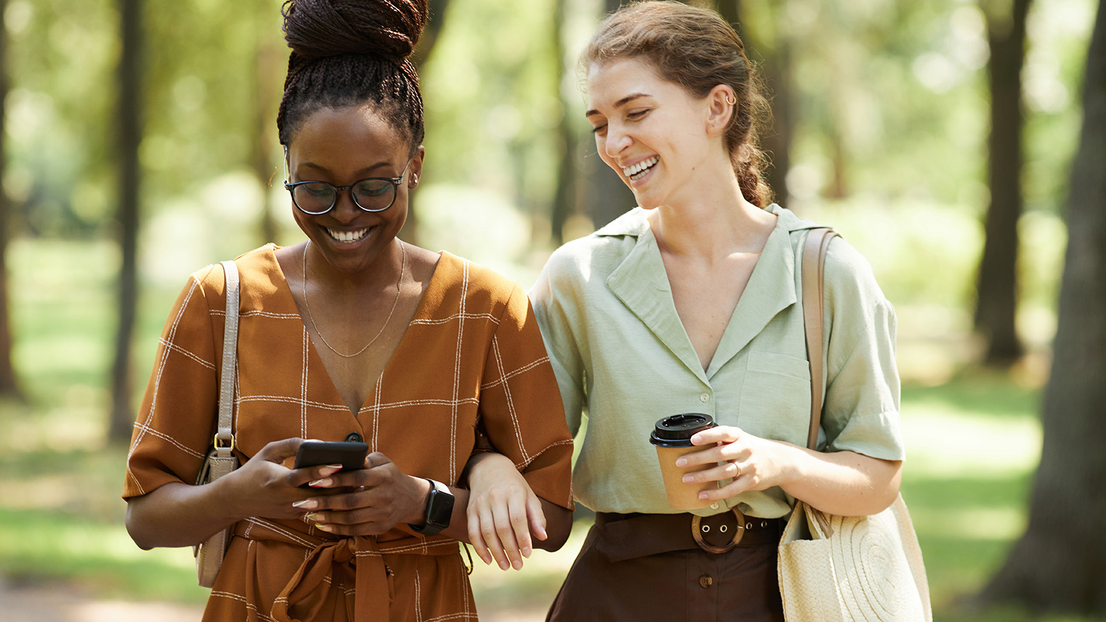 Two young women walking arm-in-arm through a park carrying phones and laughing