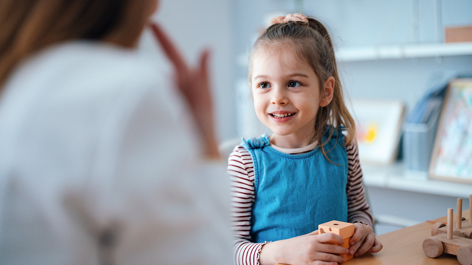 girl smiling holding wooden block toy, woman gesturing in foreground