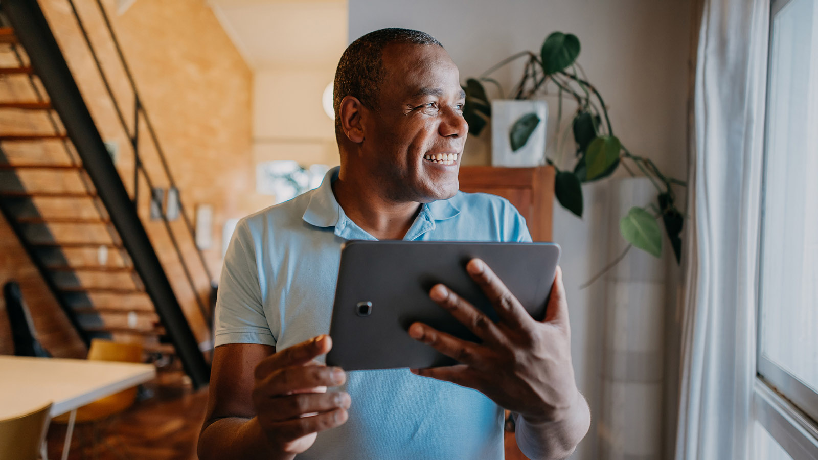 Middle aged man holding up a tablet and smiling while looking out the window