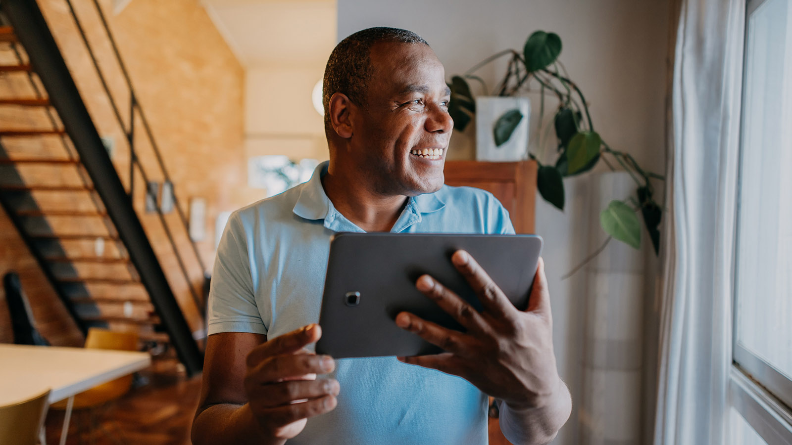Middle aged man holding up a tablet and smiling while looking out the window