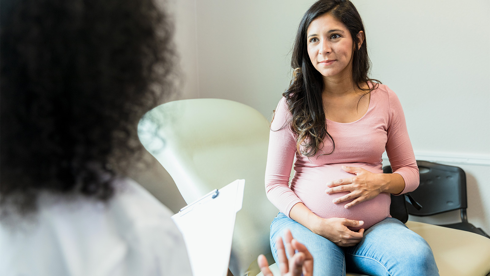 Pregnant woman sitting on table in a doctor's office with her hands on her belly, talking to doctor