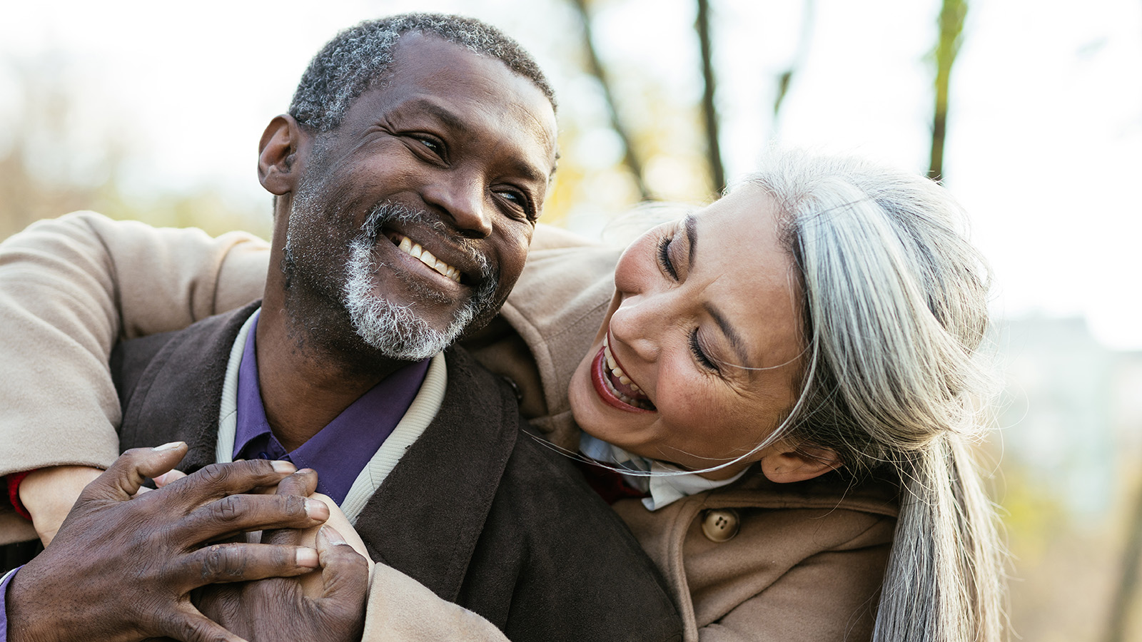 Older woman hugging an older man from behind. Both are smiling and laughing and in the outdoors.