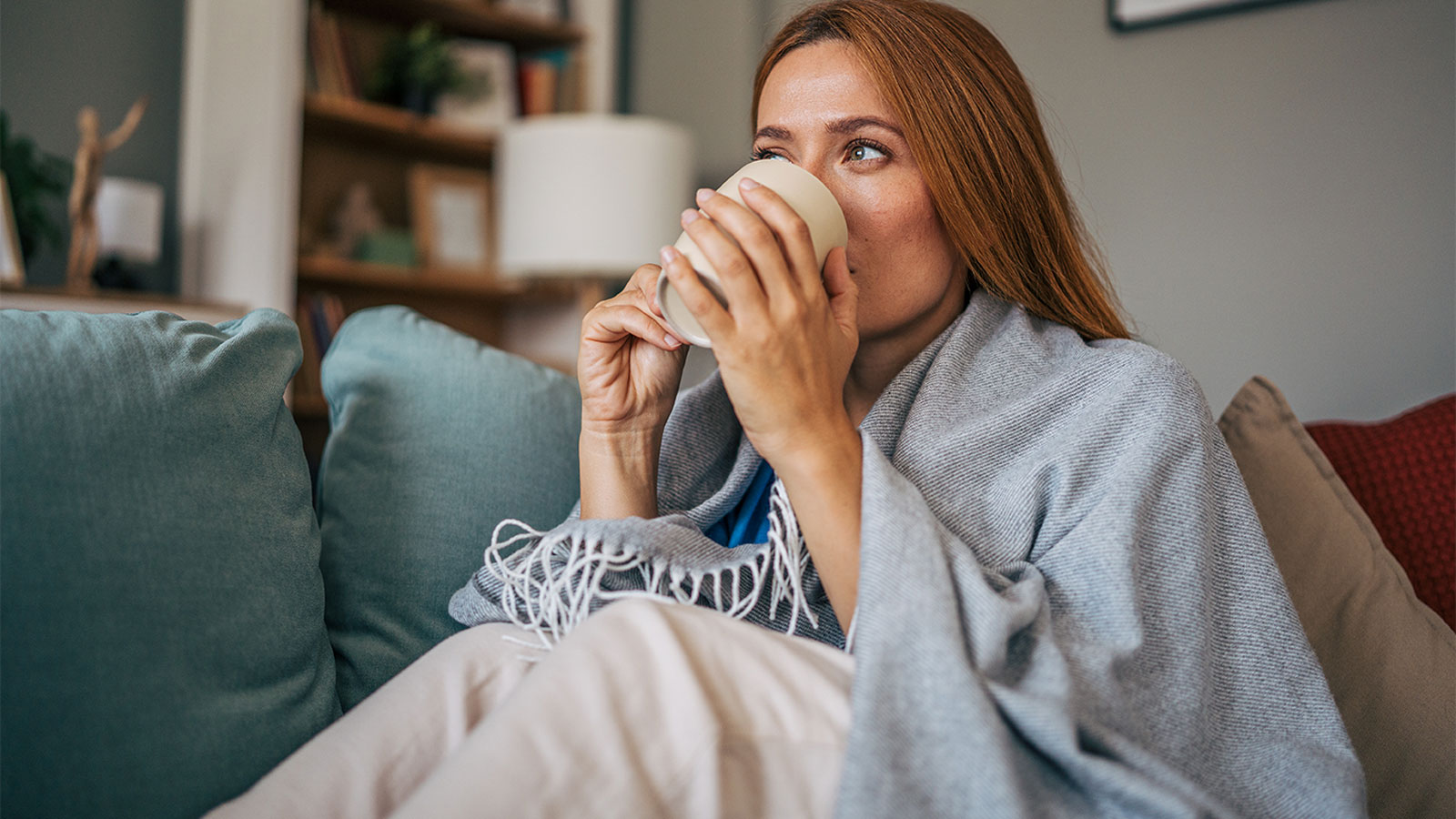 Woman sitting on couch wrapped in a blanket and drinking from a mug
