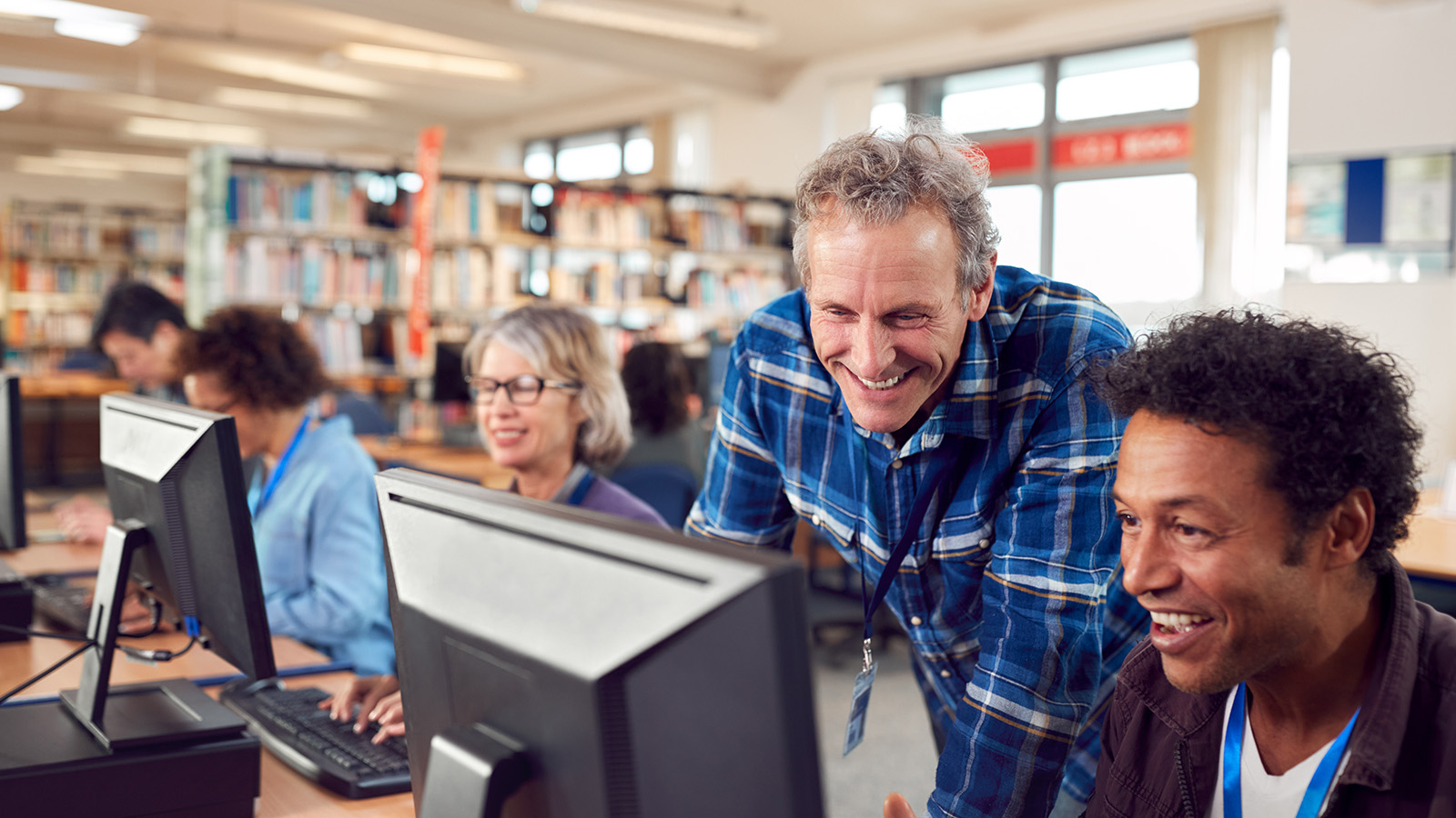 several people in a work setting at computers with one older man looking over the shoulder of another manat his screen