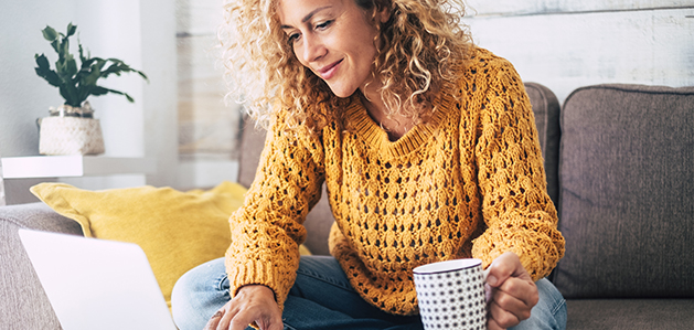Adult woman in a yellow sweater holds a cup of coffee while browsing on her laptop.