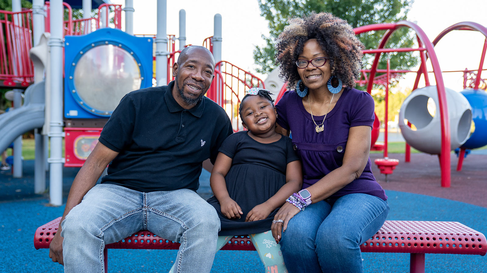Father, mother, and child pose on a colorful bench in front of playground equipment