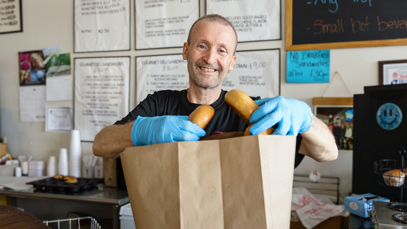 Male Virtua Heart Care patient, smiling from behind a counter, wears gloves while putting bagels into a paper bag